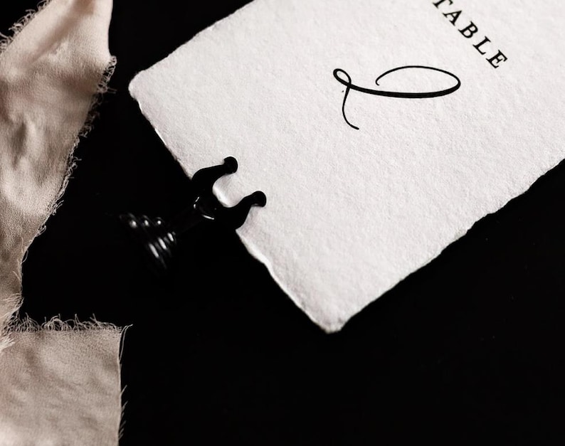 May include: A black metal place card holder gripping a white paper card with the word "TABLE" and a decorative script initial. A piece of beige fabric is in the background. The background is black.