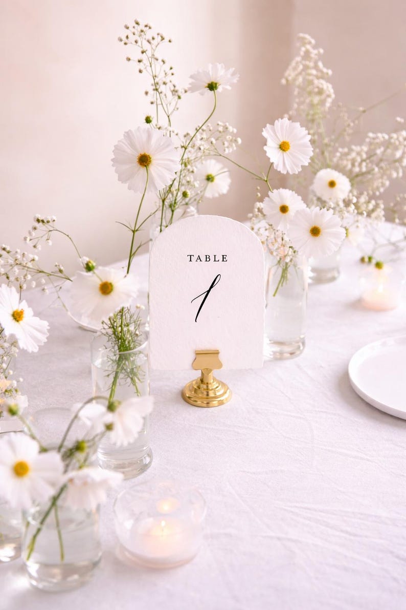 May include: A table setting with white cosmos flowers and baby's breath in small glass vases. A white card with the word "TABLE" and the number "1" is held by a gold clip. A candle in a glass holder is also on the table.