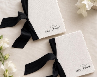 May include: Two rectangular vow books with textured white paper and deckled edges. Each book is bound with a black silk ribbon tied in a bow. One book features the black printed text "HER Vows" and the other features "HIS Vows" in script.