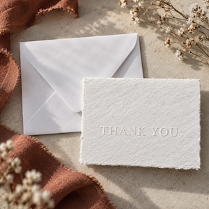 May include: A white rectangular card with a deckled edge and the words THANK YOU embossed in the center, accompanied by a matching white envelope. The card features a textured paper surface and is positioned next to a piece of rust-colored fabric.