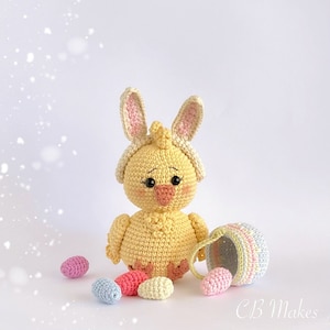 Puede incluir: Un pollito amarillo tejido a crochet con una diadema de orejas de conejo rosa y blanca. El pollito sostiene una pequeña cesta y está rodeado de huevos de Pascua de colores pastel.