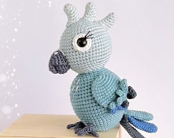 May include: A crocheted blue and gray parrot with black eyes and a white beak. The parrot is sitting on a wooden surface.