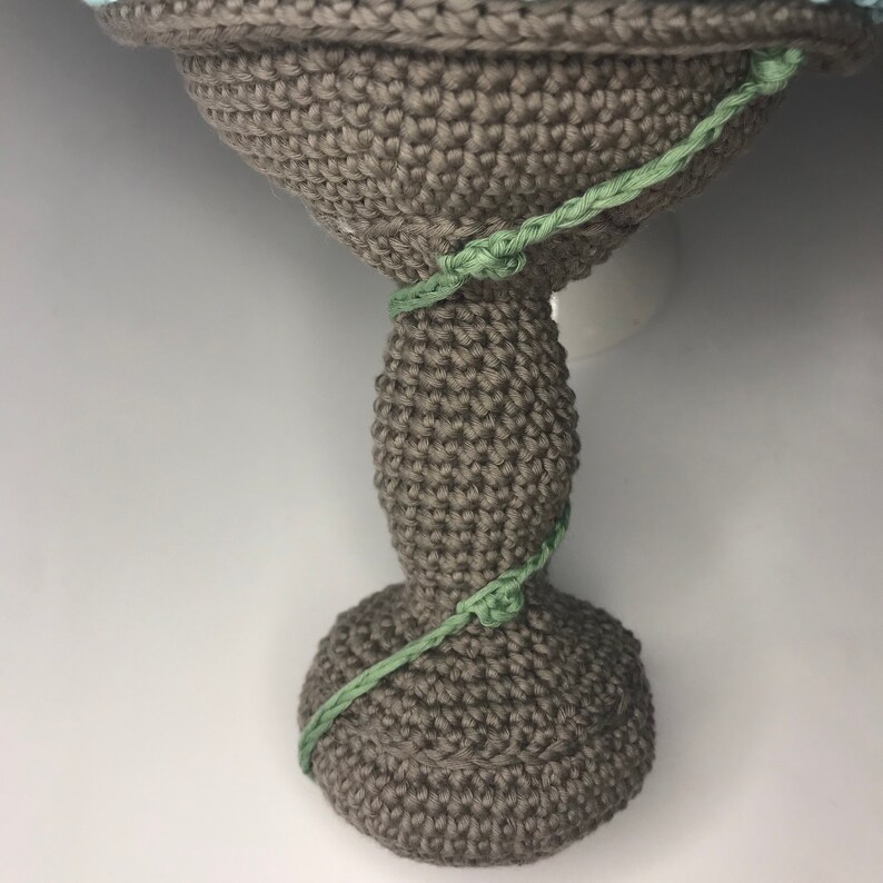 May include: A crocheted gray vase with a green crocheted vine wrapped around it.