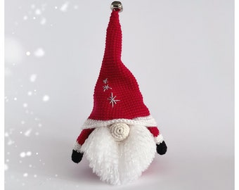 Pode incluir: Um gnomo de crochê vermelho com barba branca e um chapéu vermelho com um sino de prata no topo. O chapéu tem estrelas brancas bordadas. O gnomo está em pé sobre um fundo branco.