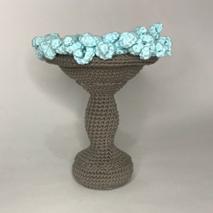 May include: A crocheted gray pedestal with a light blue crocheted top. The pedestal is shaped like a vase with a wide base and a narrow neck.