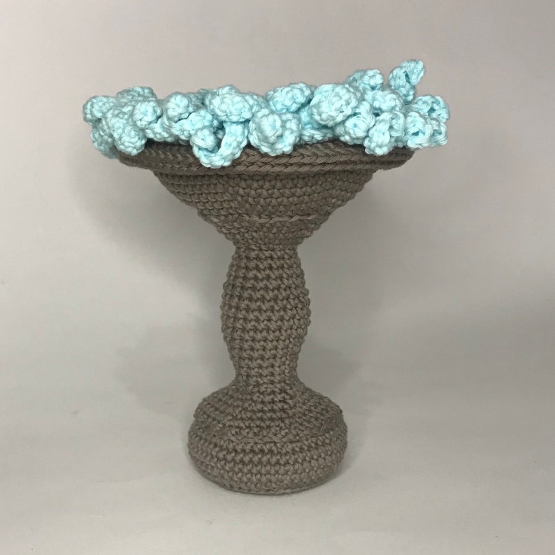 May include: A crocheted gray pedestal with a light blue crocheted top. The pedestal is shaped like a vase with a wide base and a narrow neck.