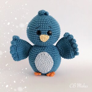 May include: A blue crocheted bird with a light blue belly, orange feet, and black eyes. The bird has its wings outstretched.