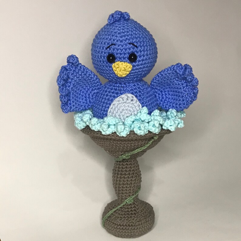 May include: A blue crocheted bird sitting in a brown crocheted bird bath with blue and white crocheted flowers. The bird bath is on a brown crocheted stand.