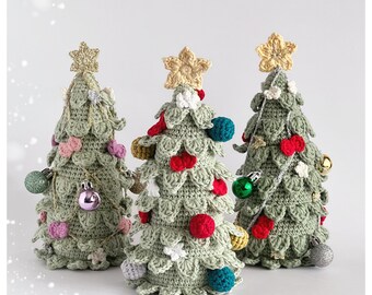 Op de afbeelding: Drie gehaakte kerstboom met verschillende gekleurde ornamenten. De bomen zijn groen en hebben een ster bovenop. De ornamenten zijn rood, groen, roze, wit en goud.