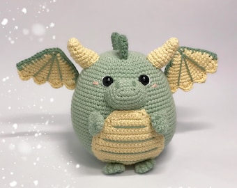 May include: A crocheted green and yellow dragon plush toy with black eyes and a small pink nose. The dragon has wings and is standing on a white surface.