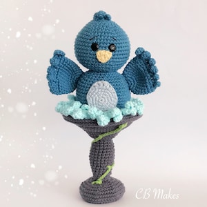 May include: A crocheted blue bird with a yellow beak and black eyes perched on a gray bird bath with green vines. The bird bath is on a white background.