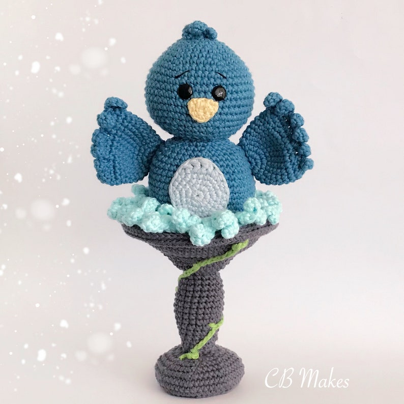 May include: A crocheted blue bird with a yellow beak and black eyes perched on a gray bird bath with green vines. The bird bath is on a white background.