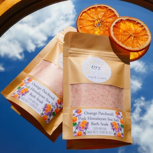 May include: Two brown paper pouches filled with pink Himalayan sea salt bath soak. The pouches have labels that read "Orange Patchouli Pink Himalayan Sea Salt Bath Soak 100% Natural Made with Essential Oils". The pouches are hanging from a gold mirror with dried orange slices in the background.