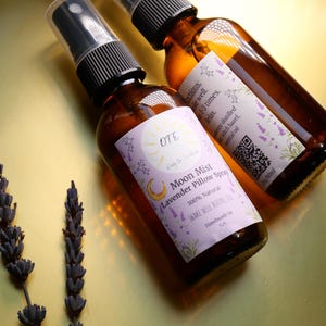 May include: Two amber glass bottles with spray nozzles, filled with a clear liquid. The bottles have labels with a crescent moon and stars, and the text "Moon Mist Lavender Pillow Spray 100% Natural Shake Well Before Use Handmade in CA".