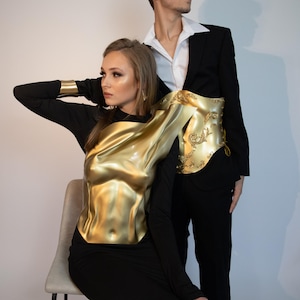 K&ouml;nnte beinhalten: Eine Frau tr&auml;gt ein goldfarbenes, geformtes Oberteil &uuml;ber einem schwarzen Kleid, und ein Mann in einem schwarzen Anzug mit wei&szlig;em Hemd. Das Oberteil hat einen metallischen Glanz und aufwendige Details. Thema: High Fashion.