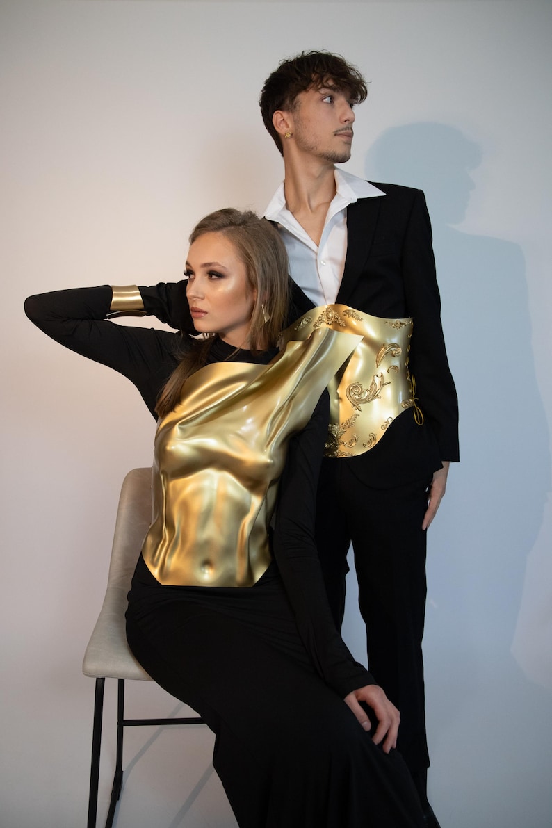 K&ouml;nnte beinhalten: Eine Frau tr&auml;gt ein goldfarbenes, geformtes Oberteil &uuml;ber einem schwarzen Kleid, und ein Mann in einem schwarzen Anzug mit wei&szlig;em Hemd. Das Oberteil hat einen metallischen Glanz und aufwendige Details. Thema: High Fashion.