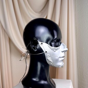 K&ouml;nnte beinhalten: Eine silberne Maske mit aufwendigen Details, pr&auml;sentiert auf einem schwarzen Schaufensterkopf. Die Maske bedeckt Nase und Untergesicht und weist komplizierte Muster und eine metallische Oberfl&auml;che auf. Sie wird mit d&uuml;nnen silbernen B&auml;ndern befestigt.