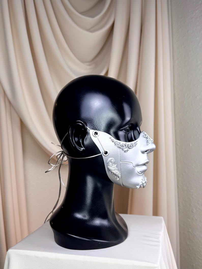 K&ouml;nnte beinhalten: Eine silberne Maske mit aufwendigen Details, pr&auml;sentiert auf einem schwarzen Schaufensterkopf. Die Maske bedeckt Nase und Untergesicht und weist komplizierte Muster und eine metallische Oberfl&auml;che auf. Sie wird mit d&uuml;nnen silbernen B&auml;ndern befestigt.