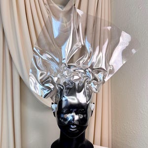 May include: A black mannequin head with a silver, sculptural headpiece. The headpiece appears to be made of a reflective, crinkled material. The mannequin has small, sparkly earrings and is set against a neutral background.