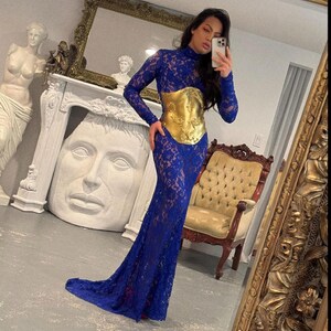 K&ouml;nnte beinhalten: Ein langes, k&ouml;nigsblaues Spitzenkleid mit langen &Auml;rmeln und hohem Ausschnitt. Das Kleid hat einen goldfarbenen G&uuml;rtel im Korsettstil. Das Kleid ist bodenlang mit einer leichten Schleppe. Das Bild wird vor einem Spiegel aufgenommen.