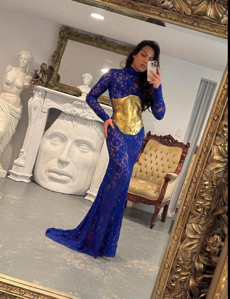 K&ouml;nnte beinhalten: Ein langes, k&ouml;nigsblaues Spitzenkleid mit langen &Auml;rmeln und hohem Ausschnitt. Das Kleid hat einen goldfarbenen G&uuml;rtel im Korsettstil. Das Kleid ist bodenlang mit einer leichten Schleppe. Das Bild wird vor einem Spiegel aufgenommen.