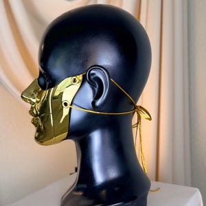 K&ouml;nnte beinhalten: Eine goldfarbene Maske mit reflektierendem Finish, die Nase und Mund bedeckt, wird auf einem schwarzen Schaufensterkopf pr&auml;sentiert. Die Maske wird mit einer goldenen Schnur befestigt, die am Hinterkopf gebunden wird. Der Hintergrund ist ein neutrales Beige.