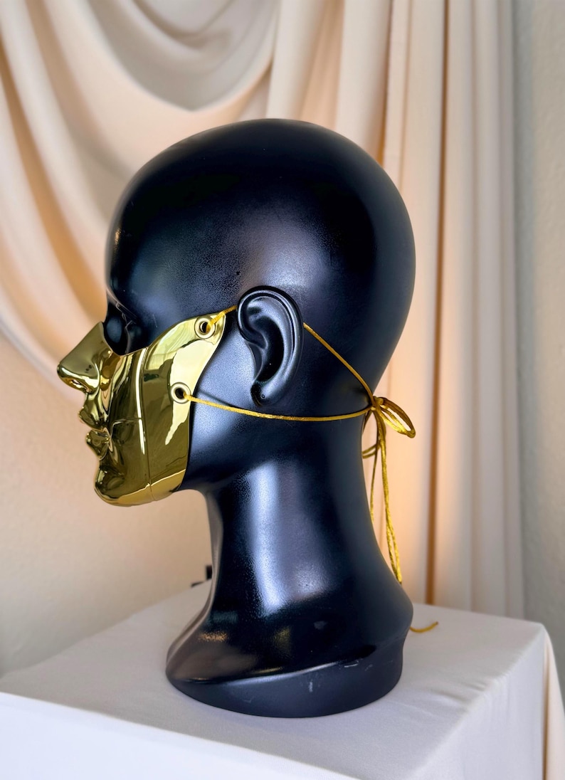 K&ouml;nnte beinhalten: Eine goldfarbene Maske mit reflektierendem Finish, die Nase und Mund bedeckt, wird auf einem schwarzen Schaufensterkopf pr&auml;sentiert. Die Maske wird mit einer goldenen Schnur befestigt, die am Hinterkopf gebunden wird. Der Hintergrund ist ein neutrales Beige.