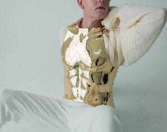 May include: A gold-colored, metallic chest plate with a sculpted, muscular design. The chest plate is worn over a cream-colored, long-sleeved shirt. The person is wearing white pants and is seated against a white background.