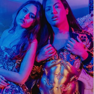 K&ouml;nnte beinhalten: Zwei Personen posieren in einem Foto, eine tr&auml;gt eine gemusterte Jacke mit goldenen Akzenten, die andere ein tr&auml;gerloses Kleid mit Gesichtsdesign. Ein goldfarbenes, verziertes K&ouml;rperteil ist der Fokus. Der Hintergrund ist in Blau und Rosa beleuchtet.