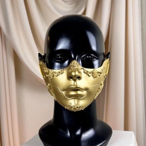 K&ouml;nnte beinhalten: Eine goldfarbene Maske mit aufwendigen Details, pr&auml;sentiert auf einem schwarzen Schaufensterkopf. Die Maske bedeckt Nase und Mund, mit dekorativen Verzierungen an den R&auml;ndern und einer glatten, metallischen Oberfl&auml;che. Der Hintergrund ist ein neutrales Beige.