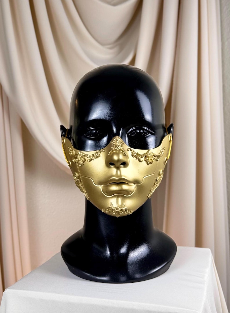 K&ouml;nnte beinhalten: Eine goldfarbene Maske mit aufwendigen Details, pr&auml;sentiert auf einem schwarzen Schaufensterkopf. Die Maske bedeckt Nase und Mund, mit dekorativen Verzierungen an den R&auml;ndern und einer glatten, metallischen Oberfl&auml;che. Der Hintergrund ist ein neutrales Beige.