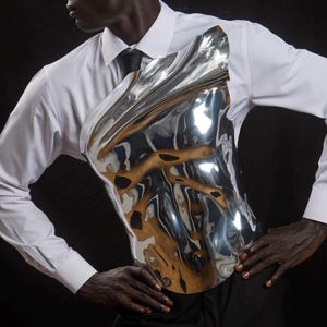 May include: A silver-colored, sculpted chest piece worn over a white dress shirt and black tie. The chest piece has a reflective, rippled surface. The person is also wearing black pants. The background is black.