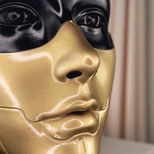 K&ouml;nnte beinhalten: Nahaufnahme eines Schaufensterpuppenkopfes mit zweifarbigem Design. Der obere Teil, einschlie&szlig;lich der Augen, ist schwarz, w&auml;hrend das untere Gesicht, die Nase und die Lippen goldfarben sind. Die Schaufensterpuppe hat eine glatte, reflektierende Oberfl&auml;che.