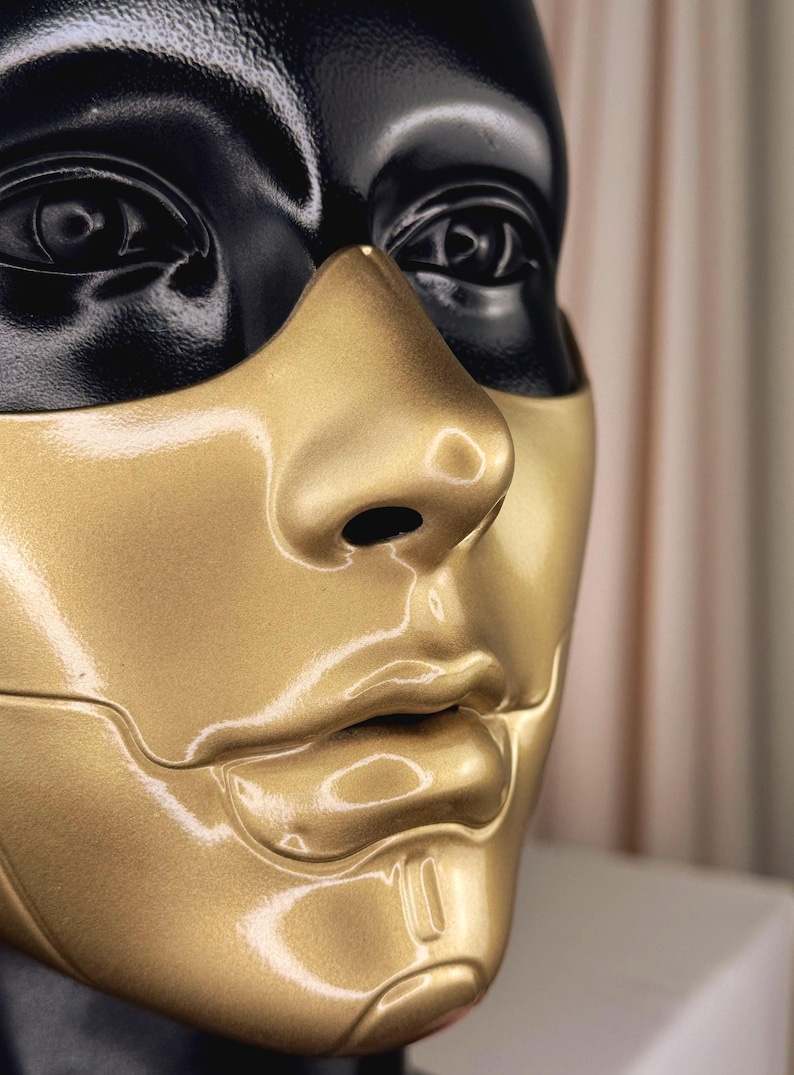 K&ouml;nnte beinhalten: Nahaufnahme eines Schaufensterpuppenkopfes mit zweifarbigem Design. Der obere Teil, einschlie&szlig;lich der Augen, ist schwarz, w&auml;hrend das untere Gesicht, die Nase und die Lippen goldfarben sind. Die Schaufensterpuppe hat eine glatte, reflektierende Oberfl&auml;che.
