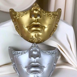K&ouml;nnte beinhalten: Zwei dekorative venezianische Masken, eine goldene und eine silberne, werden gezeigt. Die Masken weisen ein stilisiertes Gesichtsdesign mit markanten Lippen und Nase auf, verziert mit aufwendigen, wirbelnden Verzierungen. Die Masken sind wahrscheinlich f&uuml;r Kost&uuml;m- oder Dekorationszwecke.