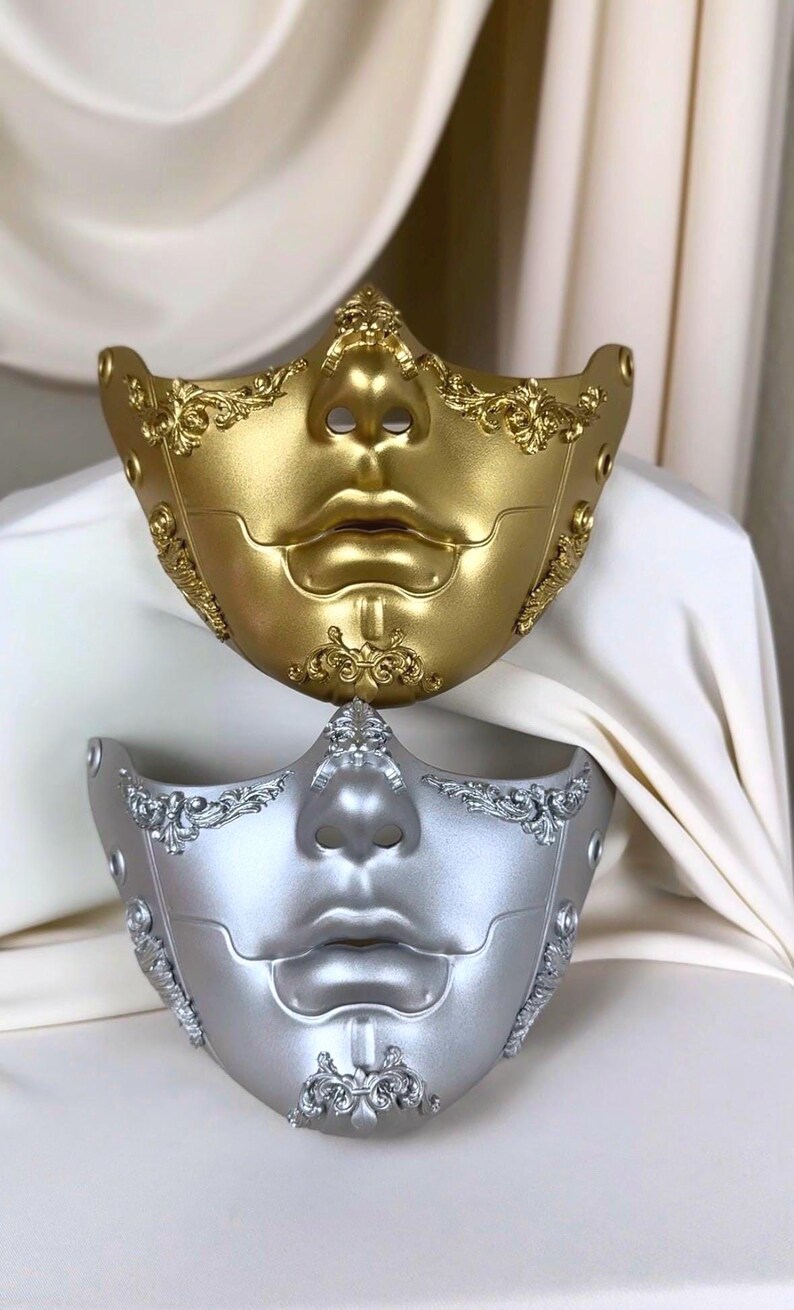 K&ouml;nnte beinhalten: Zwei dekorative venezianische Masken, eine goldene und eine silberne, werden gezeigt. Die Masken weisen ein stilisiertes Gesichtsdesign mit markanten Lippen und Nase auf, verziert mit aufwendigen, wirbelnden Verzierungen. Die Masken sind wahrscheinlich f&uuml;r Kost&uuml;m- oder Dekorationszwecke.