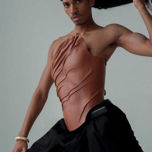Könnte beinhalten: Ein braunes Leder-Oberteil mit einem einzigartigen, geformten Design. Das Oberteil hat asymmetrische Drapierungen und eine figurbetonte Silhouette. Das Model trägt eine schwarze Hose und ein goldenes Armband. Das Oberteil ist ein Statement-Piece.
