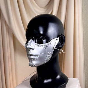 K&ouml;nnte beinhalten: Silberne und wei&szlig;e Halbmaske mit aufwendigen Details, pr&auml;sentiert auf einem schwarzen Schaufensterkopf. Die Maske hat eine metallische Oberfl&auml;che und komplizierte Designs um Nase und Wangen, mit Ohrschlaufen zum Tragen.