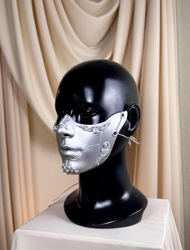 K&ouml;nnte beinhalten: Silberne und wei&szlig;e Halbmaske mit aufwendigen Details, pr&auml;sentiert auf einem schwarzen Schaufensterkopf. Die Maske hat eine metallische Oberfl&auml;che und komplizierte Designs um Nase und Wangen, mit Ohrschlaufen zum Tragen.