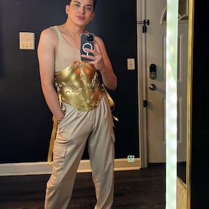 K&ouml;nnte beinhalten: Ein goldfarbenes, verziertes Oberteil im Korsettstil mit aufwendigen Details wird mit beigefarbenen Cargohosen kombiniert. Das Outfit umfasst ein beiges Mesh-Top und eine goldfarbene Sch&auml;rpe. Die Person h&auml;lt ein Telefon.