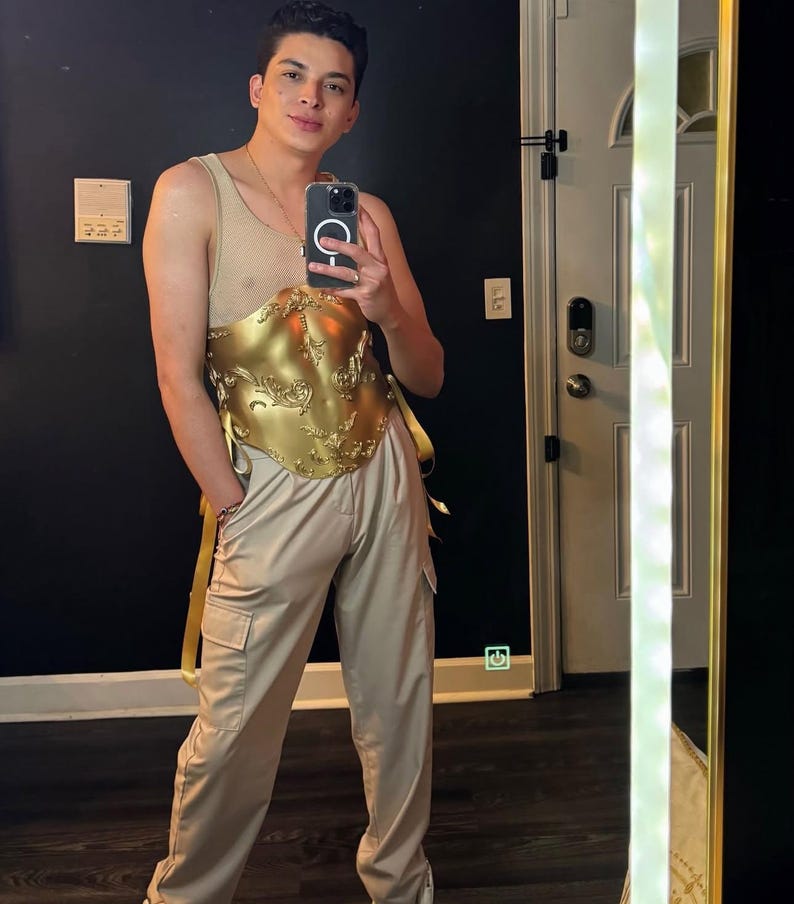 K&ouml;nnte beinhalten: Ein goldfarbenes, verziertes Oberteil im Korsettstil mit aufwendigen Details wird mit beigefarbenen Cargohosen kombiniert. Das Outfit umfasst ein beiges Mesh-Top und eine goldfarbene Sch&auml;rpe. Die Person h&auml;lt ein Telefon.
