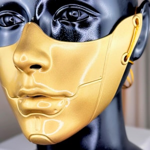 K&ouml;nnte beinhalten: Eine goldfarbene Maske mit metallischem Finish, die das untere Gesicht und die Nase bedeckt. Die Maske ist an einem schwarzen Schaufensterpuppenkopf befestigt. Die Maske hat ein futuristisches Design mit sichtbaren Linien und einer glatten Oberfl&auml;che. Die Maske ist mit einer gelben Schnur befestigt.