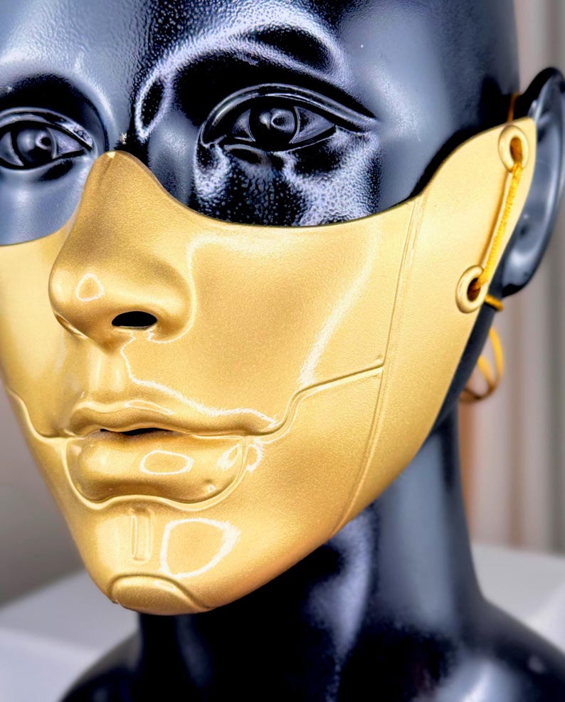 K&ouml;nnte beinhalten: Eine goldfarbene Maske mit metallischem Finish, die das untere Gesicht und die Nase bedeckt. Die Maske ist an einem schwarzen Schaufensterpuppenkopf befestigt. Die Maske hat ein futuristisches Design mit sichtbaren Linien und einer glatten Oberfl&auml;che. Die Maske ist mit einer gelben Schnur befestigt.