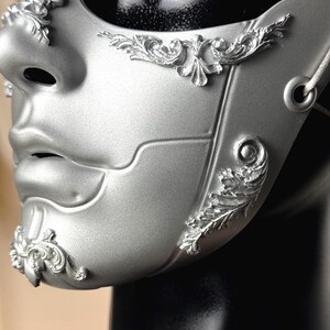 K&ouml;nnte beinhalten: Eine silberne Maske f&uuml;r einen Maskenball mit erhabenen floralen Designs. Die Maske bedeckt Nase und Mund und hat ein detailliertes, geformtes Aussehen. Die Maske ist an einem schwarzen Schaufensterpuppenkopf befestigt.