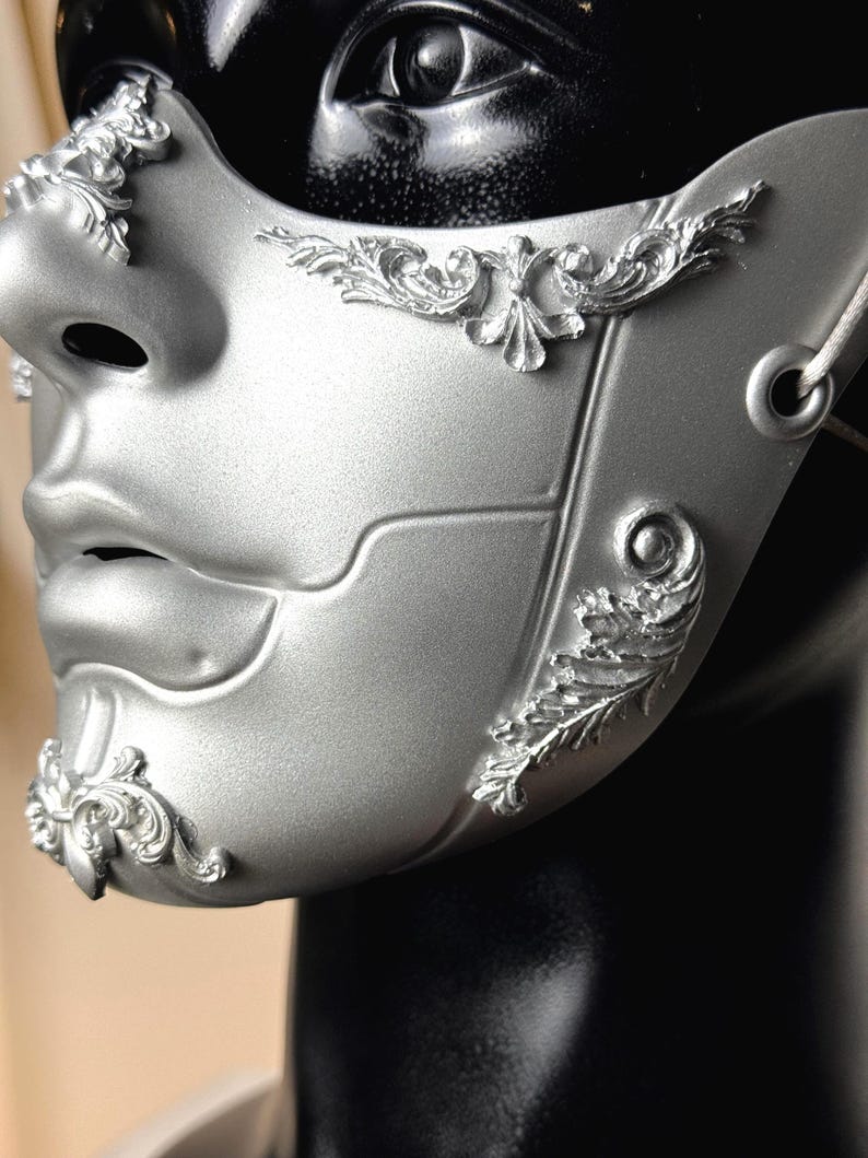K&ouml;nnte beinhalten: Eine silberne Maske f&uuml;r einen Maskenball mit erhabenen floralen Designs. Die Maske bedeckt Nase und Mund und hat ein detailliertes, geformtes Aussehen. Die Maske ist an einem schwarzen Schaufensterpuppenkopf befestigt.