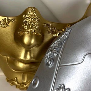 K&ouml;nnte beinhalten: Nahaufnahme von zwei dekorativen Masken. Eine ist goldfarben mit aufwendigen floralen Details, die andere ist silberfarben mit &auml;hnlichen Verzierungen. Die Masken haben eine glatte, metallische Oberfl&auml;che und dienen wahrscheinlich Kost&uuml;m- oder Dekorationszwecken.