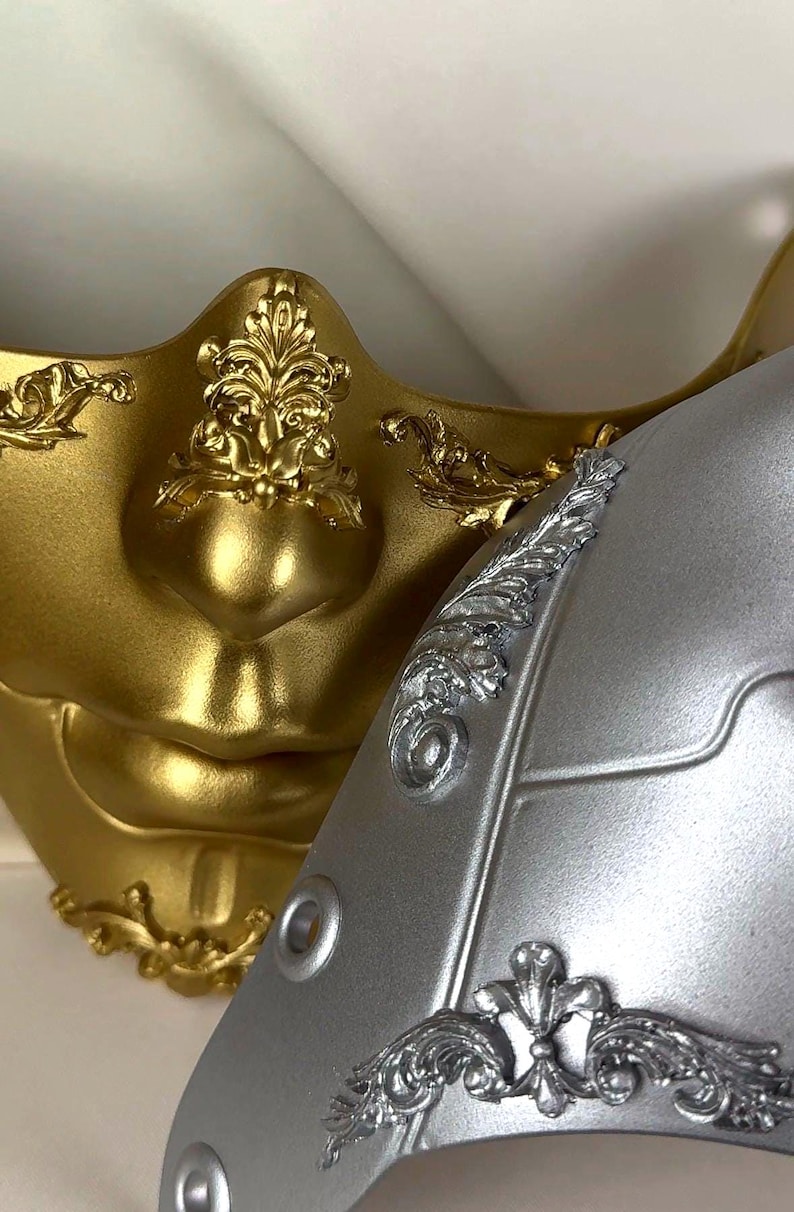 K&ouml;nnte beinhalten: Nahaufnahme von zwei dekorativen Masken. Eine ist goldfarben mit aufwendigen floralen Details, die andere ist silberfarben mit &auml;hnlichen Verzierungen. Die Masken haben eine glatte, metallische Oberfl&auml;che und dienen wahrscheinlich Kost&uuml;m- oder Dekorationszwecken.