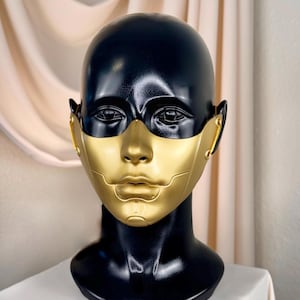 May include: A black mannequin head with a gold-colored face mask. The mask covers the nose and mouth, with detailed features. The mannequin is on a white surface, with a neutral background.