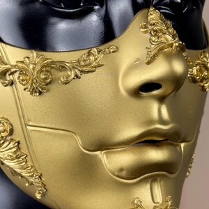 K&ouml;nnte beinhalten: Eine goldfarbene Maske mit aufwendigen, erhabenen floralen Designs. Die Maske bedeckt Nase und Mund, mit einer glatten, reflektierenden Oberfl&auml;che. Die Maske ist vor einem schwarzen Hintergrund platziert.