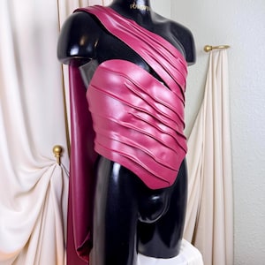 May include: A burgundy-colored costume with a draped shoulder piece and a pleated bodice. The costume is displayed on a black mannequin. A matching cape hangs from the shoulder. The mannequin is on a white pedestal.